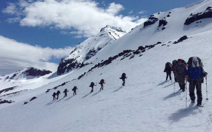 A line of people wearing snow gear, carrying backpacks and using trekking poles, hike through the snow of a mountainous landscape.